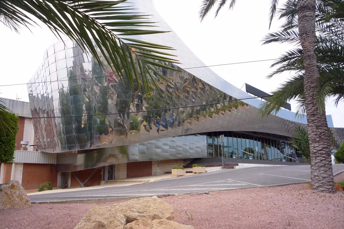 贝尼多姆宫（Benidorm Palace）的弧形转角，覆以抛光镜面钢，前方是棕榈叶与一条道路